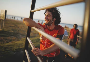 Gordijnen Portrait of a smiling young athletic male enjoying the music from his earphones resting after exercising at the outdoors gym © StratfordProductions