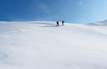 Fototapeta na wymiar Two men climbing on mountain peak on skis or splitboards at white snow and blue sky background. Ski touring sport activity