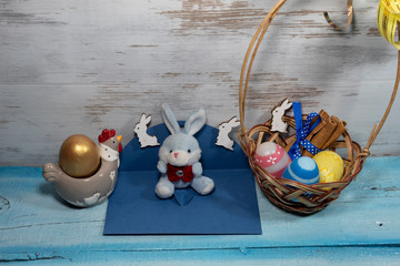 A toy rabbit sits on an open blue envelope, two hares on clothespins. Near a wicker basket with Easter eggs and a chicken with an egg.