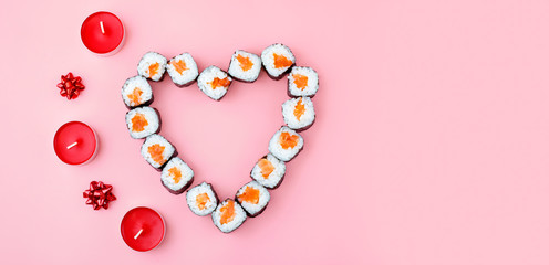 Salmon sushi rolls laid out in the shape of a heart, next to red candles and bowson a pink background. The concept of Japanese cuisine for Valentine's Day, greeting card, banner. Copy space