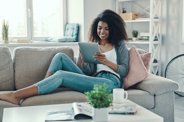 Relaxed young African woman using digital tablet and smiling while resting at home