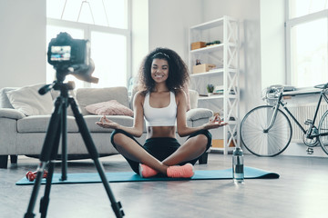 Fototapeta na wymiar Attractive young African woman sitting in yoga position and smiling while making social media video