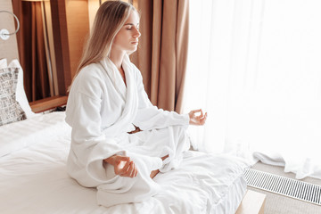 Obraz na płótnie Canvas Peaceful calm beautiful young caucasian woman blonde in white coat is meditating with closed eyes while sitting in lotus position on her bed on sunny summer morning. Healthy start day concept