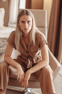 Confident beautiful positive young business lady in luxurious brown overalls in comfortable leather armchair in her hotel room. Fashion concept. Female model poses for glossy magazine. Style, trend