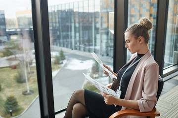 Young businesswoman sitting on chair in front of the big window and examining financial reports