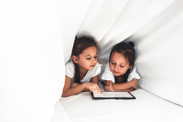 Fototapeta premium Two beautiful little sisters lying in the bed and look at the screen of a tablet, smart technology