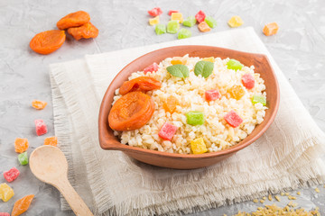 Bulgur porridge with dried apricots and candied fruits in clay bowl on a gray concrete background and linen textile. Side view, close up