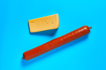 One piece of cheese in form of triangle near whole long sausage lies on blue desk on kitchen or market. Top view. Close-up