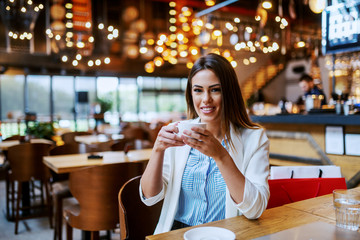 Gorgeous fashionable caucasian brunette with beautiful smile sitting in coffee shop and holding cup of fresh delicious espresso. Behind her backs on chair are shopping bags.