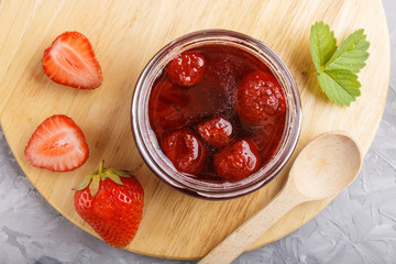 Strawberry jam in a glass jar with berries and leaves on gray concrete background, top view.