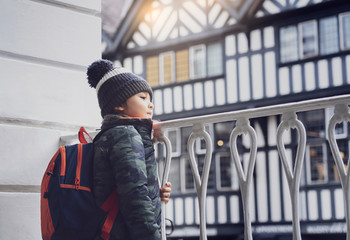 Obraz premium Candid shot of little boy looking through metal fench on street, Cropped shot of Backpack child with warm winter cloth adventure in the city, Side view portrait kid looking out with smiling face