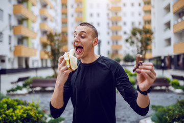 Attractive sportsman standing outdoors and choosing between candy and fresh banana. In background are buildings.