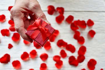 Male hand with a gift box on a background of red rose petals. St. Valentine's Day background. 