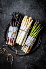 Bunches of fresh green, purple, white asparagus on vintage metal tray over dark grey rustic background. Top view, copy space