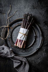 Bunch of fresh purple asparagus on vintage metal tray over dark grey rustic background. Top view, copy space