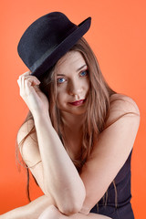 Young attractive caucasian brunette woman with bright blue eyes and dark lipstick, touches the black bowler hat, looking right to the camera. Isolated on living coral background, studio, copy space.