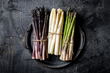 Bunches of fresh green, purple, white asparagus on vintage metal tray over dark grey rustic...