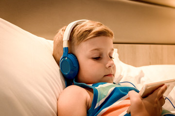 Small kid with headphones using smart phone in bedroom