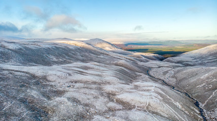 An scenic aerial view of a snowy Scottish mountain pass with stream and green forest valley and mountain range in the backgound under a majestic blue sky