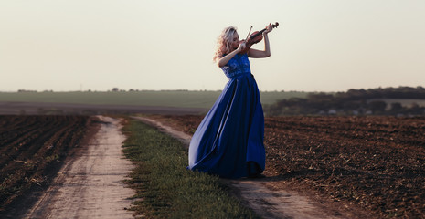 woman in long dress playing violin on background of field path with a club of dust, girl engaged in musical art, performance on nature, concept passion in music © fantom_rd