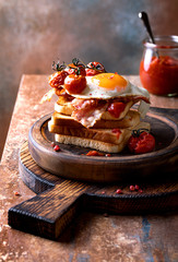 Toast with fried egg, bacon, roasted cherry tomatoes and barbecue relish on the wooden cutting board.