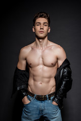 Fototapeta na wymiar sexy young man with muscular torso in biker jacket and jeans posing with hands in pockets on black background