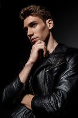 sexy young man in biker jacket touching chin and looking away on black background