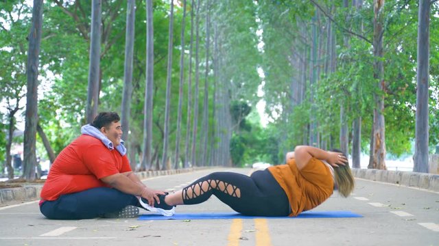 Overweight man helping his girlfriend doing sit-up exercise at the park. Shot in 4k resolution