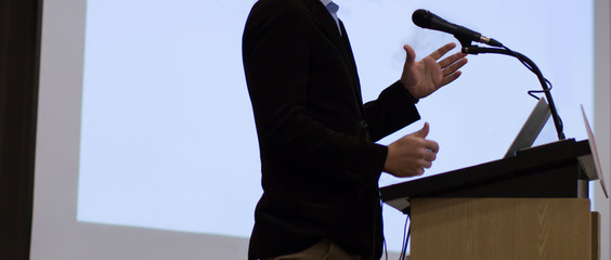 Speaker giving a talk at a corporate business conference. Audience in hall with presenter in front of presentation screen. Corporate executive giving speech during business and entrepreneur seminar. 