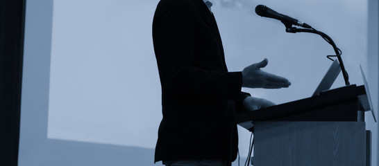 Speaker giving a talk at a corporate business conference. Audience in hall with presenter in front of presentation screen. Corporate executive giving speech during business and entrepreneur seminar. 