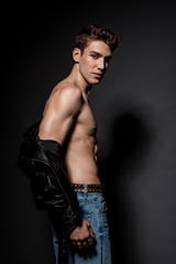 Fototapeta na wymiar side view of sexy young man with muscular torso in biker jacket and jeans on black background