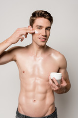 smiling sexy man with muscular torso applying face cream isolated on grey