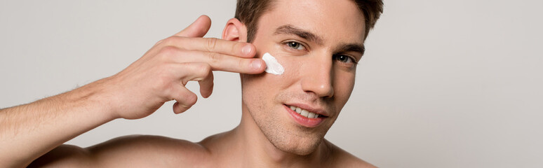 smiling sexy man with muscular torso applying face cream isolated on grey, panoramic shot