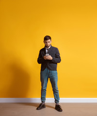 Fototapeta na wymiar All the life in gadget. Young caucasian man using smartphone, serfing, chatting, betting. Full length portrait isolated on yellow background. Concept of modern technologies, millennials, social media.