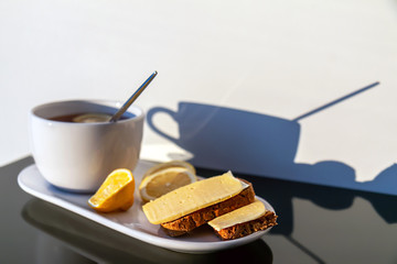 mug with black tea and lemon and ex shadow on the wall under the bright sun, breakfast with bread...