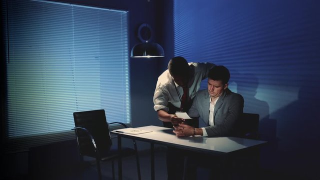 Black detective using force in questioning the accused man while showing a photo of somebody. He forces him to confess something in interrogation room.