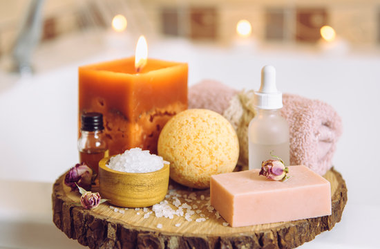 Home spa products on wooden disc tray: bar of soap, bath bomb, aroma bath salt, essential and massage oils, candle burning, rolled towel inside bathroom by tub, water running. Cozy relaxing concept.