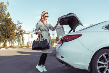 Girl in the autumn in city in parking lot, in summer by car. Opens trunk and removes and removes shopping bag. White business sedan. Parking at the mall.