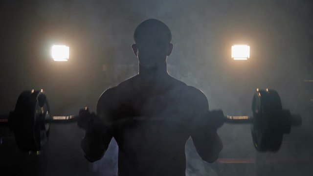 Muscular man doing crossfit training in a dark shadowy gym lifting weights holding a barbell at waist level in a health and fitness concept, movement round shot