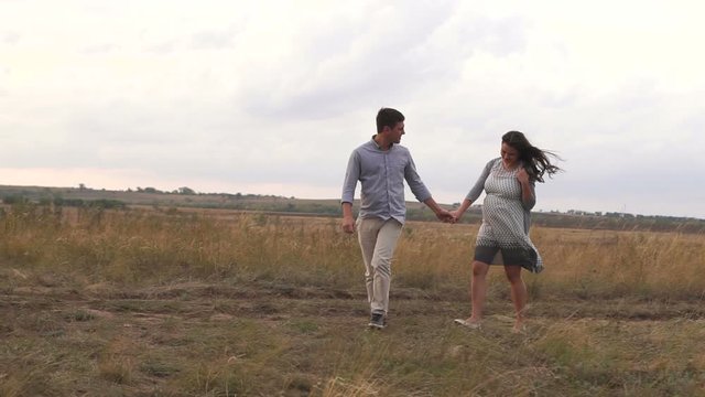 A happy pregnant family walks in a field at sunset, a man holding his wife's hand. Pregnant woman with her beloved husband in nature in windy weather.
