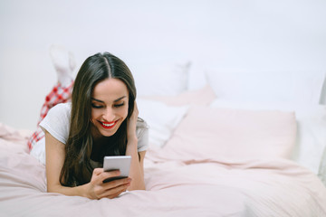 Attractive young smiling woman lying in bed in cozy light colored bedroom texting message on her mobile phone