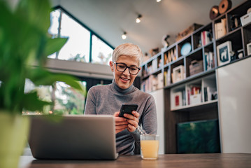 Smiling trendy senior woman using smart phone in home office, portrait.