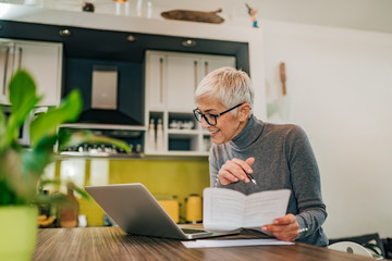 Smiling senior woman working with documents and laptop in the kitchen at modern home.