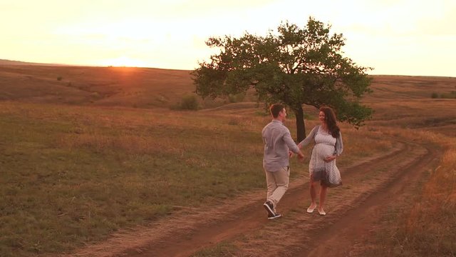 A happy pregnant woman and her husband are walking along a country road in a field at sunset in summer. Slow motion.