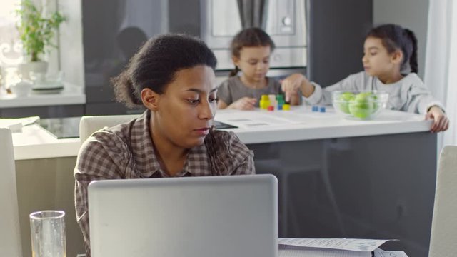 PAN of black woman working from home: she sitting at kitchen table and typing on laptop while inspecting document; preschool and primary age sisters playing with building blocks in background