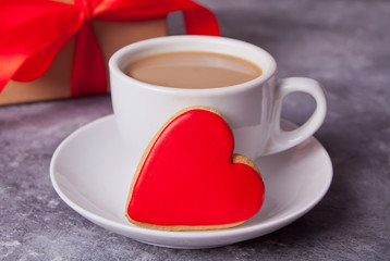 Cup of coffee and a heart shaped red cookie with gift box on the background.