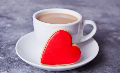 Cup of coffee and a heart shaped red cookie on the gray table.