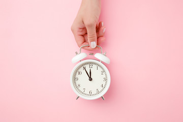 Young woman hand holding white alarm clock on light pink table background. Pastel color. Time...