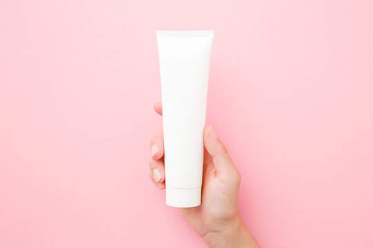 Young Woman Hand Holding And Showing White Tube On Pastel Pink Table. Care About Clean And Soft Body Skin. Daily Beauty Product. Closeup. Top Down View. Point Of View Shot.