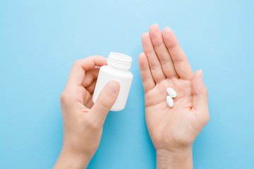 Young adult woman spilling out white pills from bottle in her hand. Receiving vitamins or medicaments.  Point of view shot. Closeup. Pastel blue table background. Top down view.